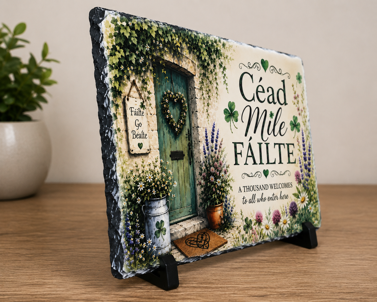 A rectangular slate sign with a rustic black edge, featuring a painted Irish welcome scene with a green door, ivy, flowers, shamrocks, and the text 'Céad Mile Fáilte' and 'A Thousand welcomes to all who enter here', displayed on a small black stand on a wooden surface.